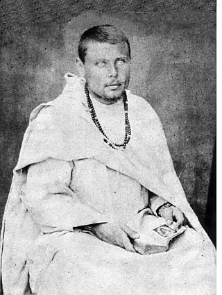 Old photograph of a bearded young European priest in a white robe and a long rosary, sitting with a book in his hands