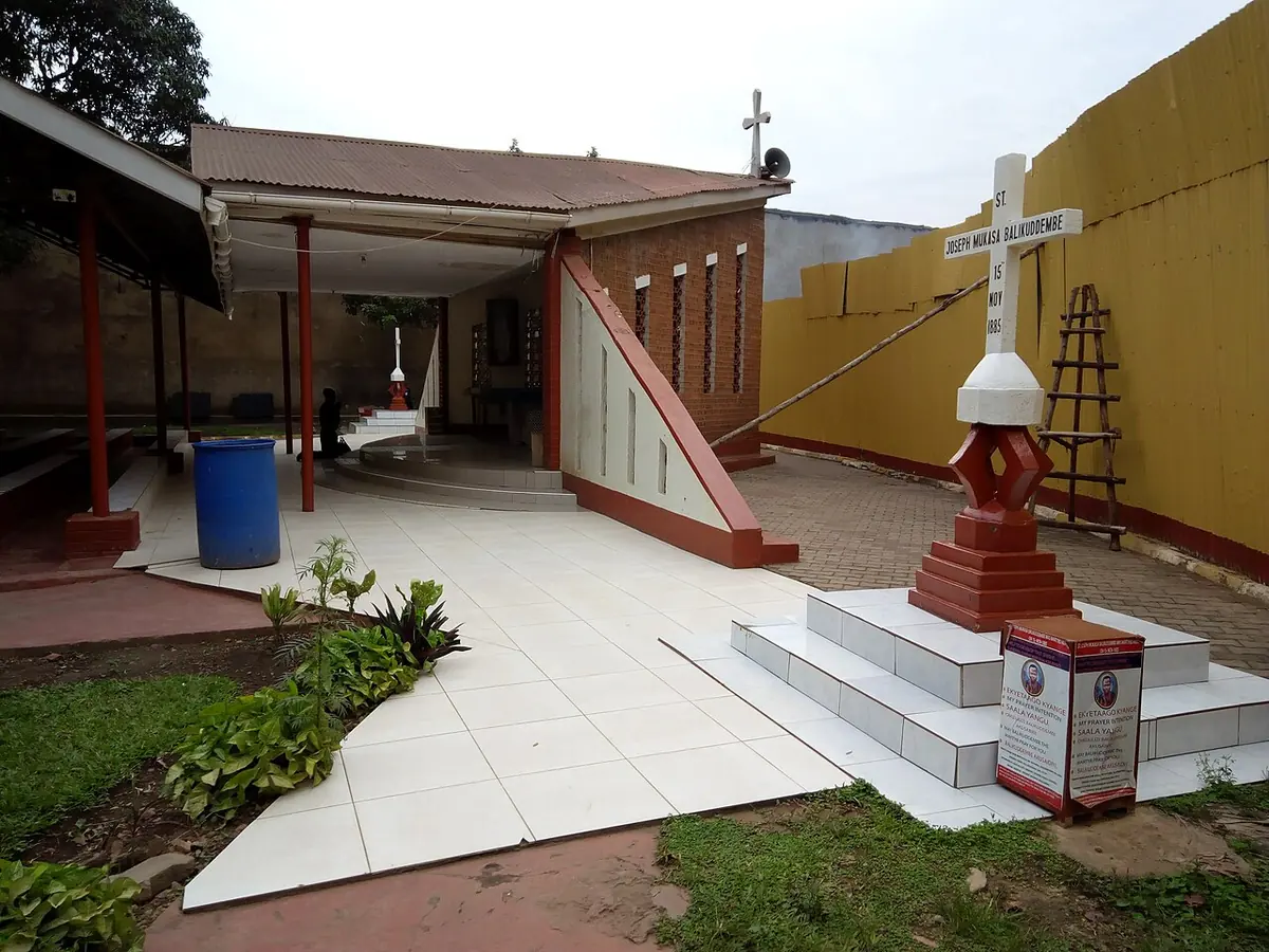A small open-sided chapel with a red roof and a white cross monument on a stepped base, inside a walled yard in a busy city