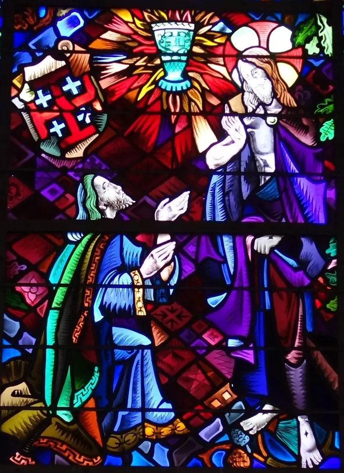 Brightly colored stained glass of a kneeling old man in a blue robe before the risen Christ, with a glowing chalice above them and a shield of red crosses