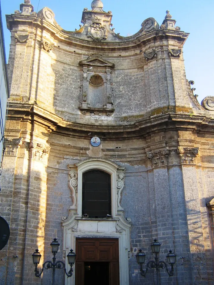 The curved Baroque stone facade of the sanctuary in Copertino, with a niche statue above the door and a wrought-iron lamp on each side