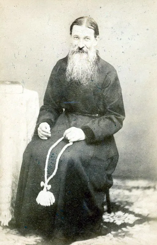 Old photograph of an elderly monk with a long white beard in a black cassock, seated with a prayer rope in his hands