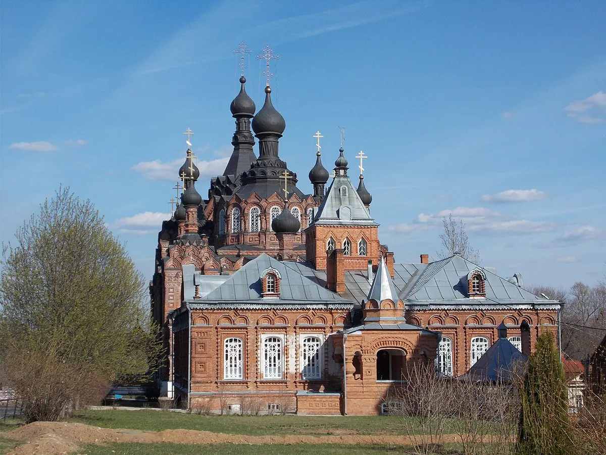 A large red-brick church with many dark onion domes and a sloping metal roof, beside a green lawn and young trees