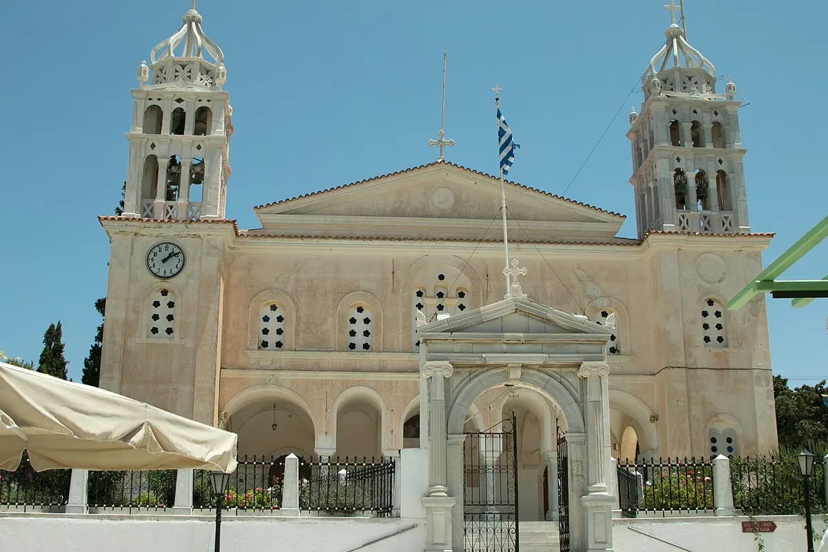 The marble facade and twin clock towers of the 19th-century Church of the Holy Trinity in Lefkes on Paros, with an arched gate in front