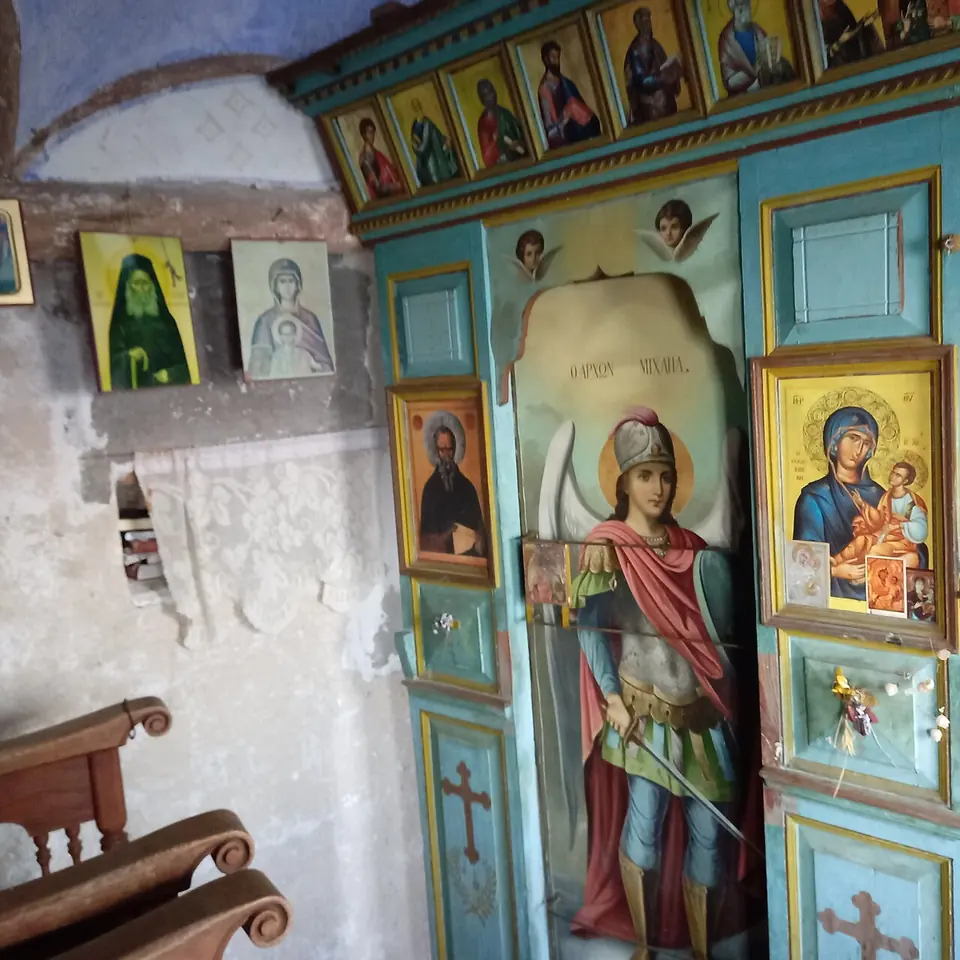 The bare interior of a hermit's cell with icons on whitewashed walls, a lit oil lamp on a small table and a wooden stall by the door