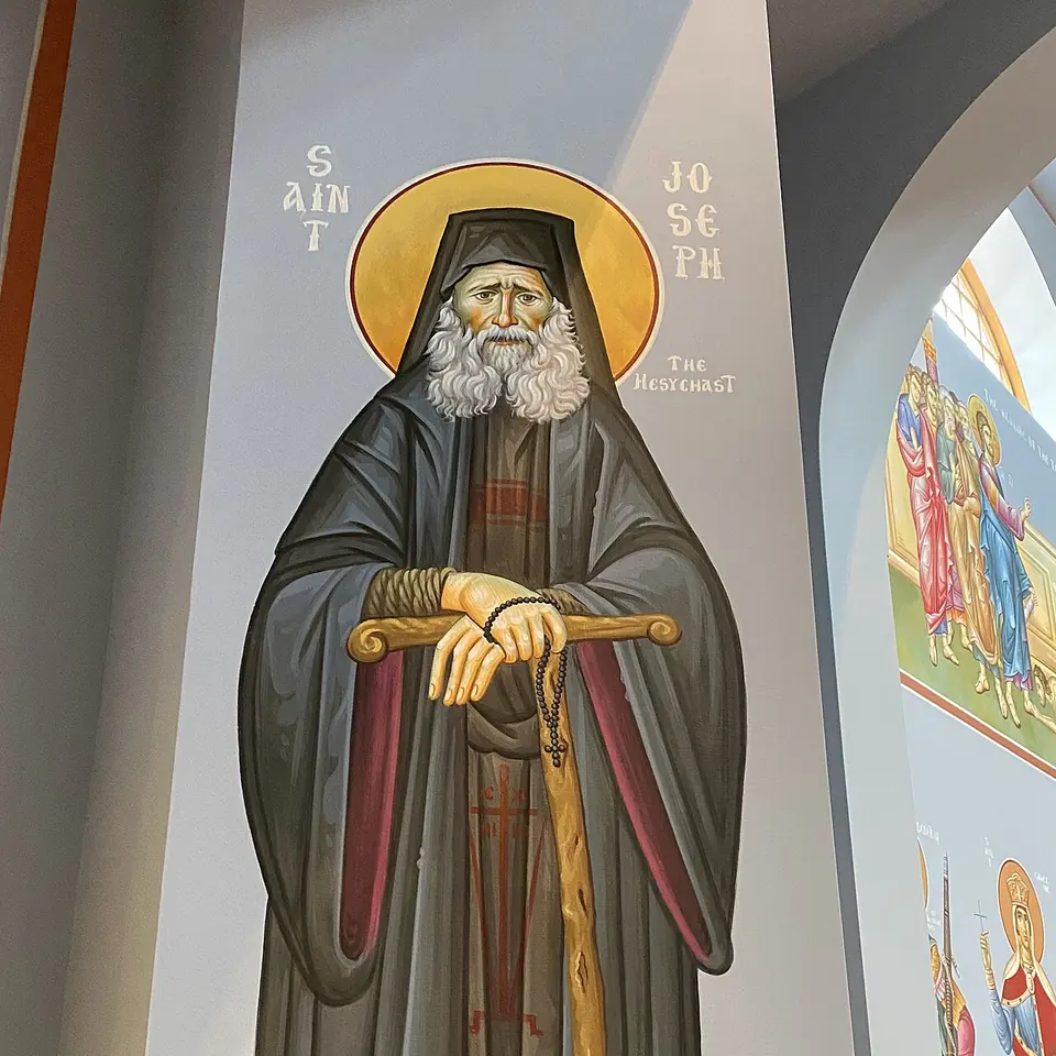 Painted wall icon of St. Joseph the Hesychast standing with a gold halo, white beard and black monastic hood, both hands resting on a T-shaped staff, a prayer rope hanging from his fingers