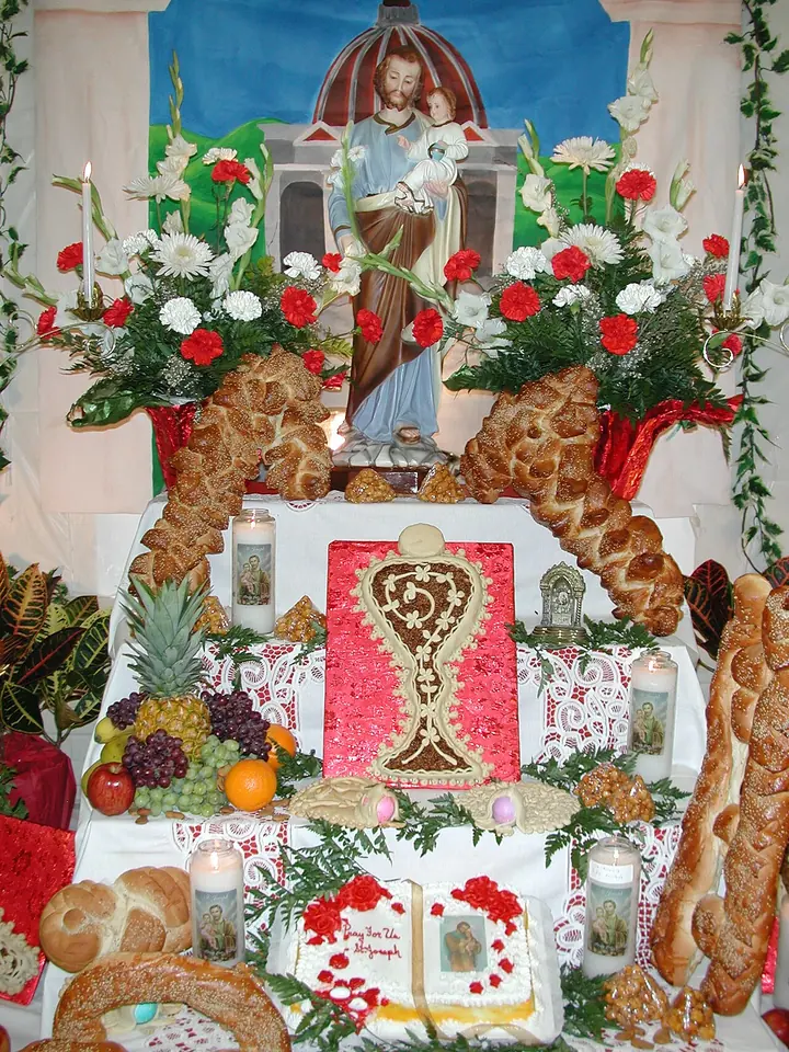 A St. Joseph's Day altar topped by a statue of Joseph holding the Child, surrounded by red and white carnations, braided loaves, fruit, candles and a large decorated bread in the shape of a monstrance