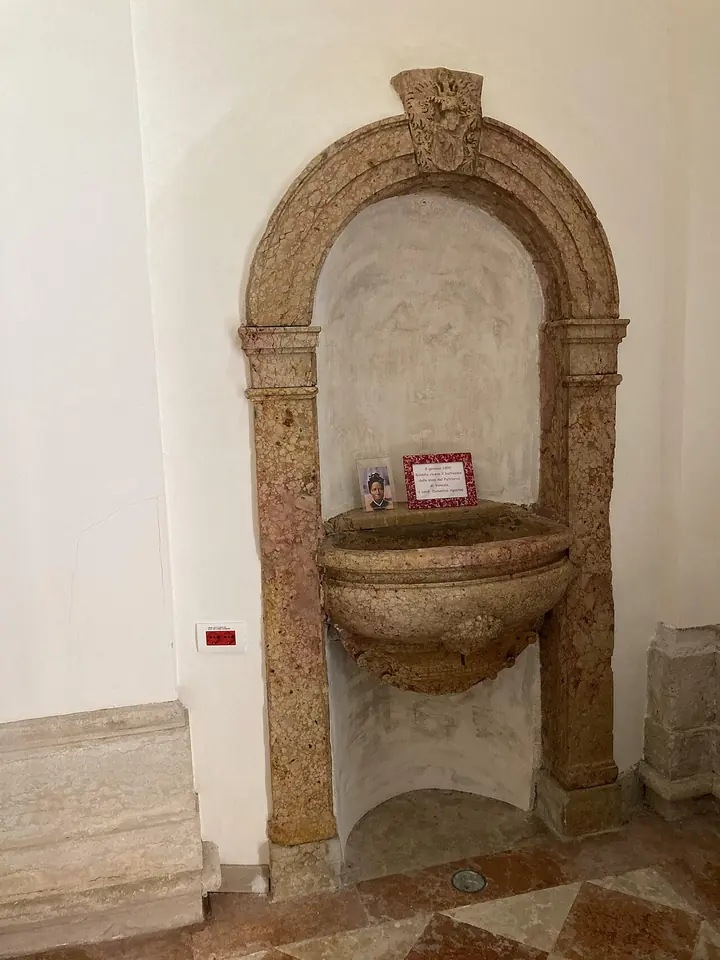 A stone baptismal font set in an arched marble niche in a white church wall, with a small picture of Bakhita placed inside