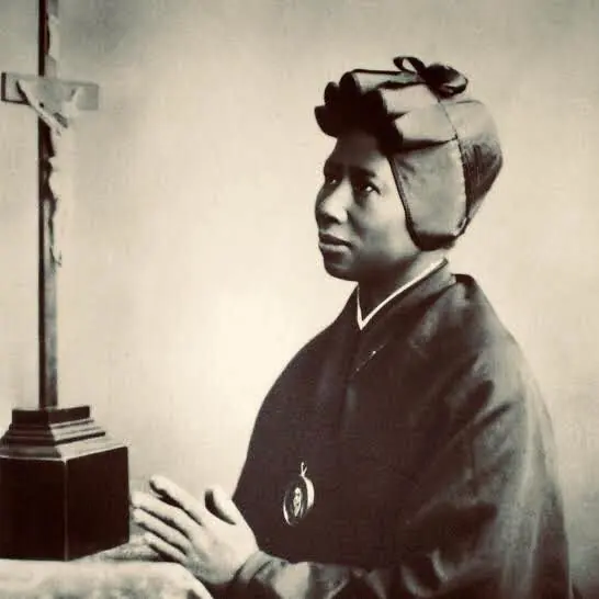 Old sepia photograph of Bakhita in her habit and bonnet, a medal at her chest, kneeling with folded hands beside a standing crucifix