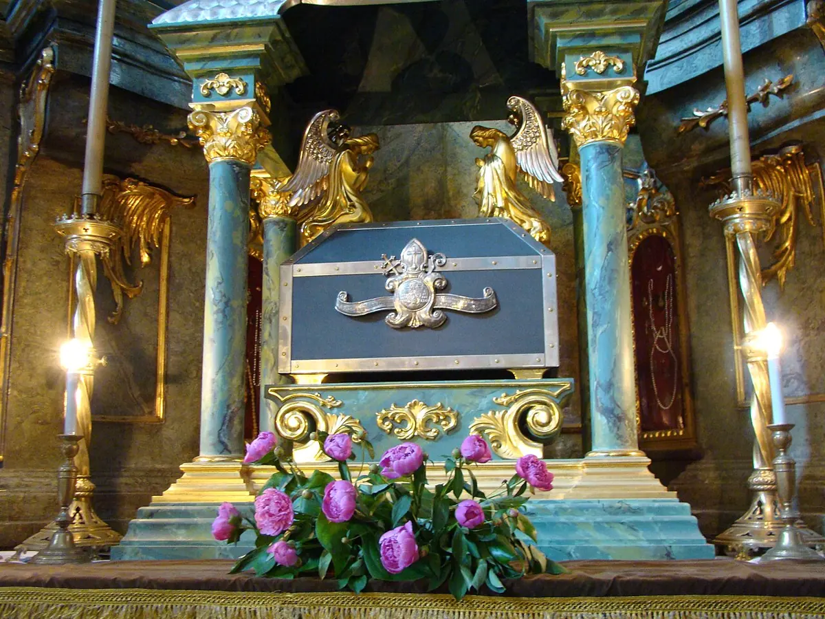 A silver-and-blue reliquary chest on a marble altar between gilded angels and candles, with pink peonies in front