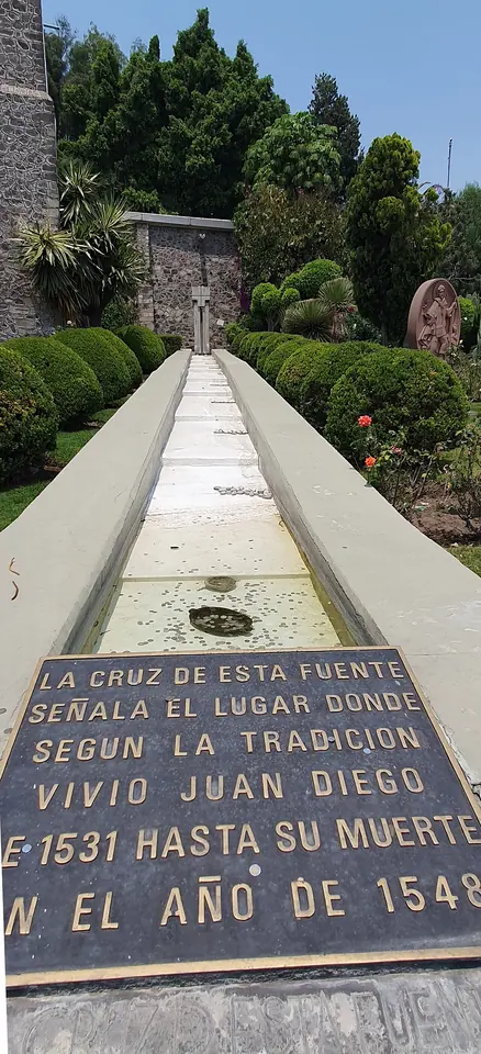 A long stone fountain in a garden with a bronze plaque in Spanish marking the place where, by tradition, Juan Diego lived from 1531 until his death in 1548