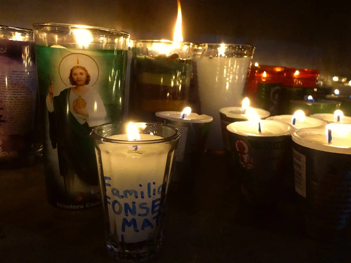 Lit glass votive candles in the dark, one printed with a picture of St. Jude in a green robe and another with a family name written on it in marker