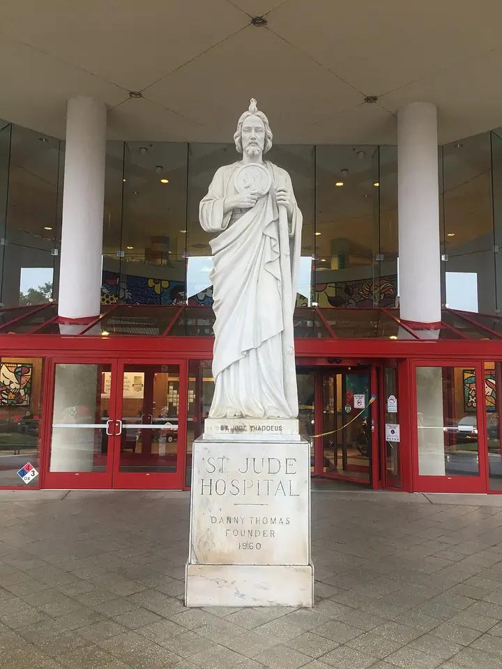 White statue of St. Jude on a pedestal inscribed "St. Jude Hospital, Danny Thomas, Founder" in front of the hospital's glass entrance