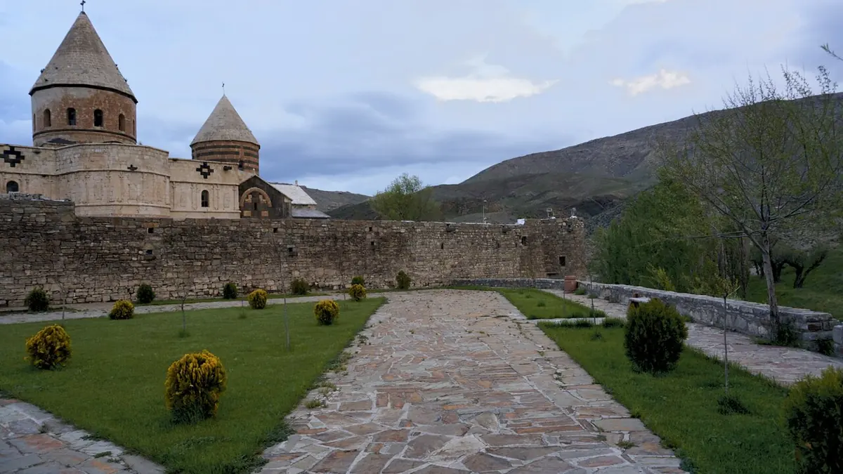 Stone Armenian church with two conical domes behind a walled garden path, with green hills beyond