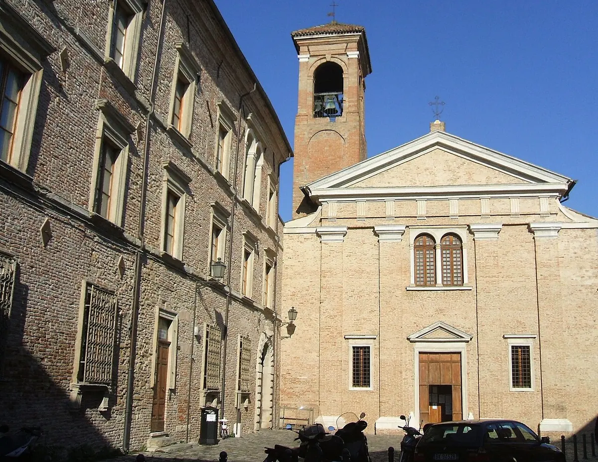A plain brick church facade with pilasters and a small bell tower beside a long monastery building, under a bright blue sky