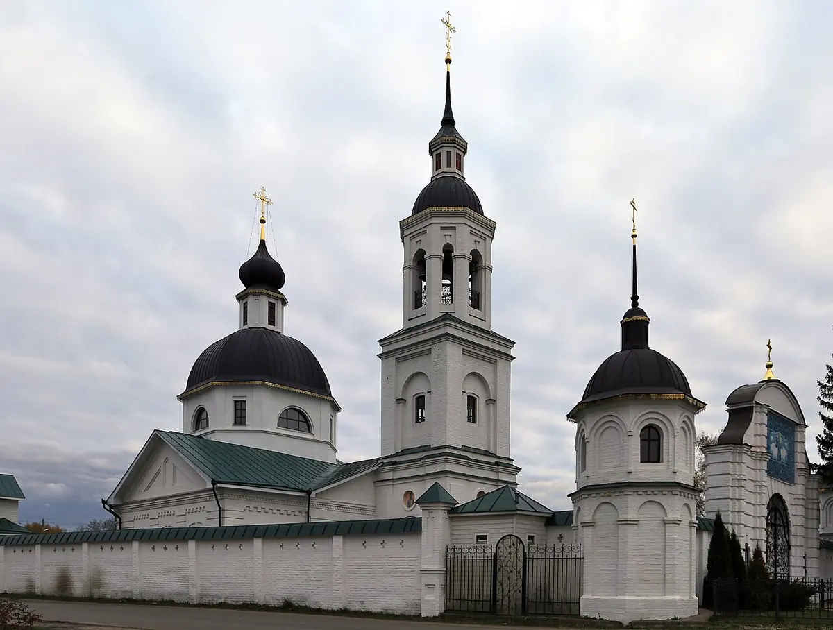 White village church with dark domes and a tall bell tower behind a white wall under a cloudy sky