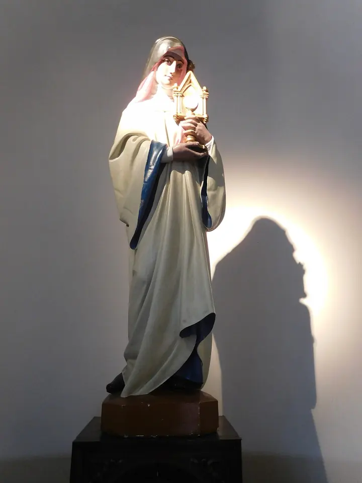 Painted statue of a nun in a white habit with a blue-lined cloak and red veil lining, holding a golden monstrance against her chest