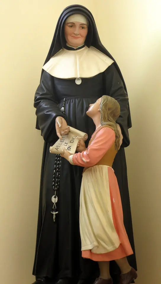 Painted statue of a nun in a black habit and white collar holding a scroll that a young girl in a pink dress and apron reads, looking up at her