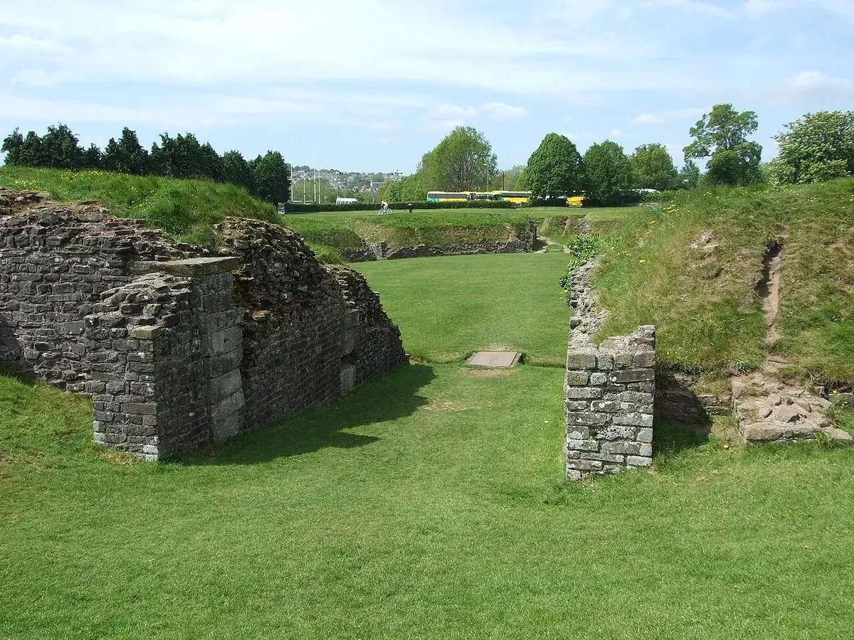 The grassy oval arena of a Roman amphitheater with low ruined stone walls and an entrance passage, under a pale sky