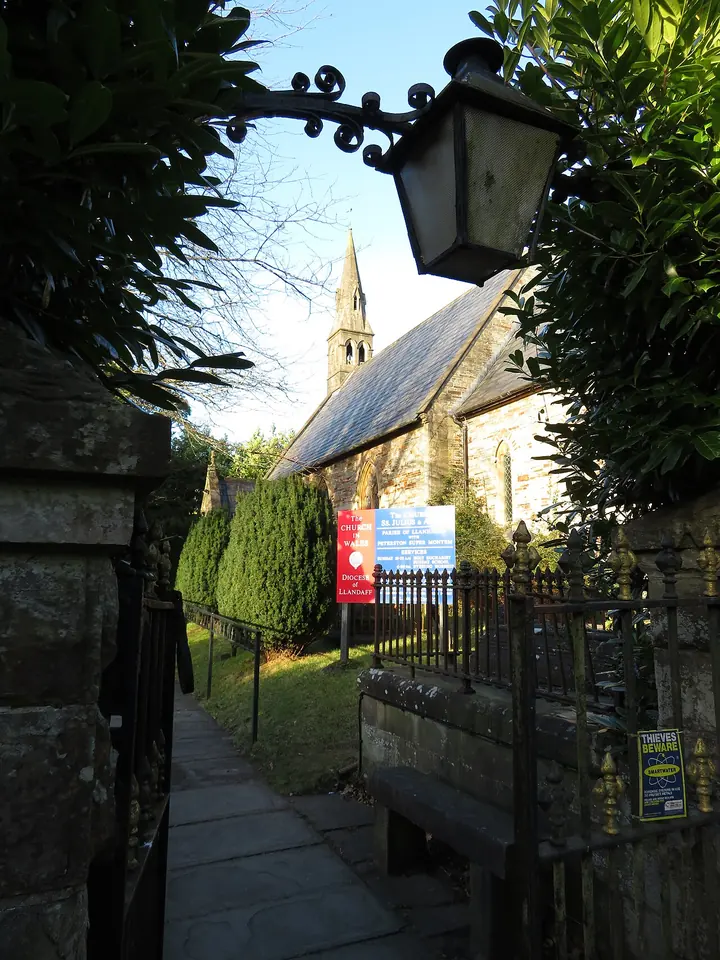 A stone church with a slender spire seen down a narrow path between a stone wall and an iron lamp bracket