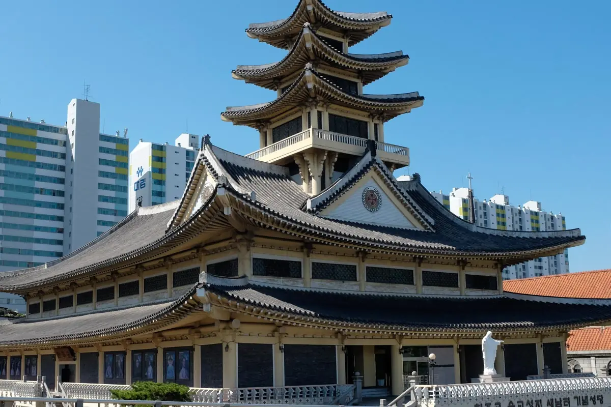 A Korean-style church with a three-tiered pagoda roof and white walls under a blue sky, with apartment towers behind
