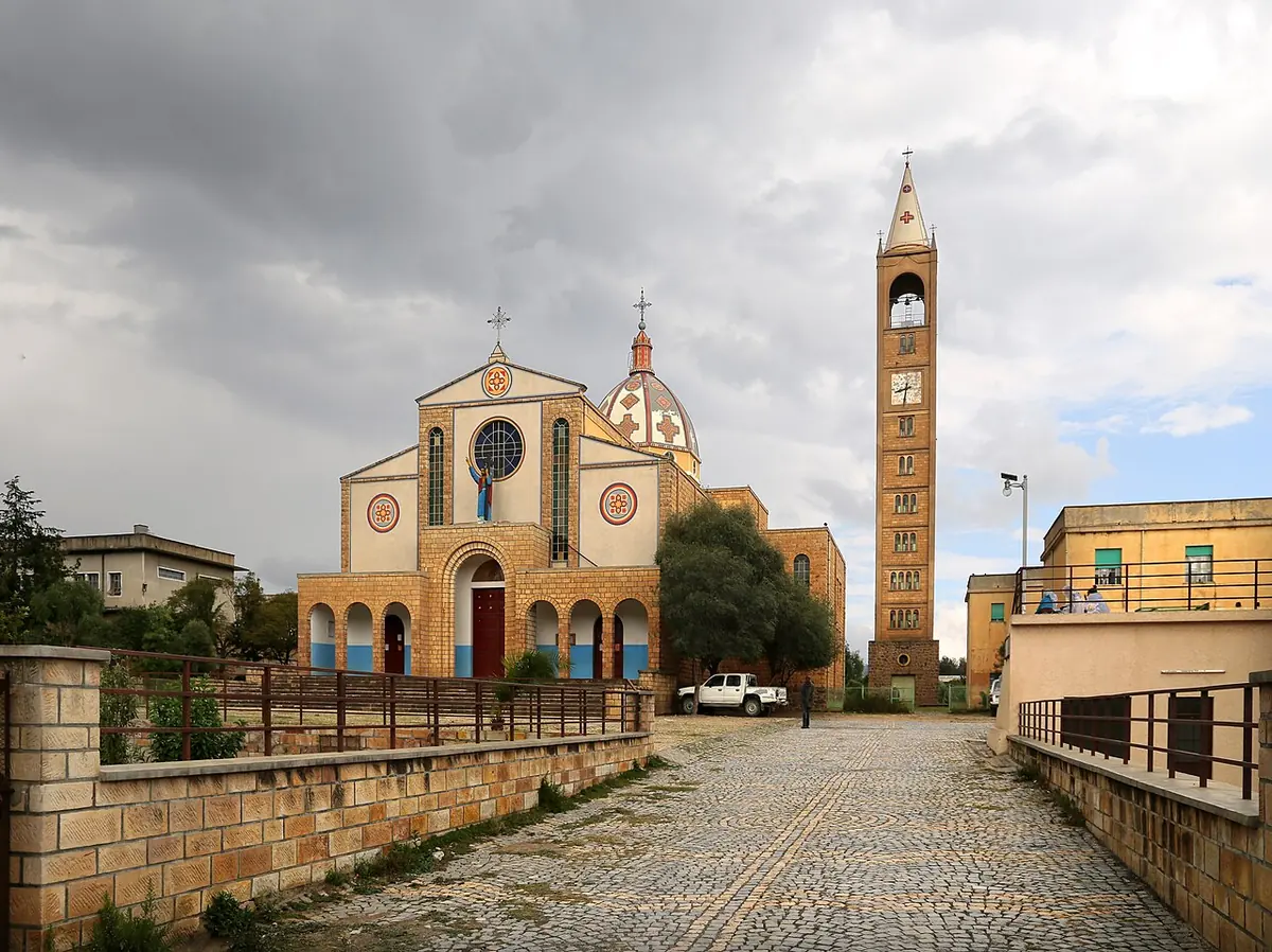 A cream and red church facade with a rose window and a tall slender bell tower at the end of a cobbled walkway, under a cloudy sky