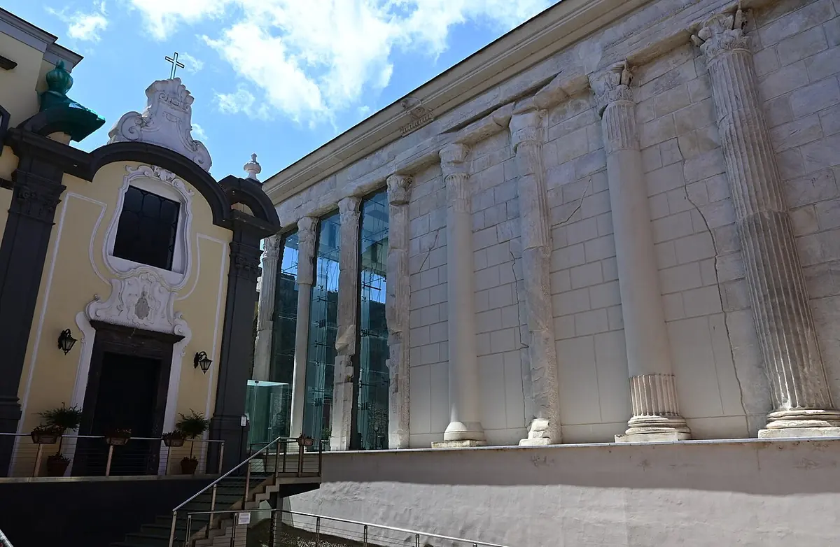 The white marble columns of an ancient Roman temple built into the side of a cathedral, next to a cream-colored church facade with a dark stone doorway