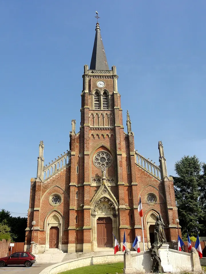 Red-brick Gothic Revival church with a tall slate spire and rose window, flags in front, in the town of Saint-Just-en-Chaussée