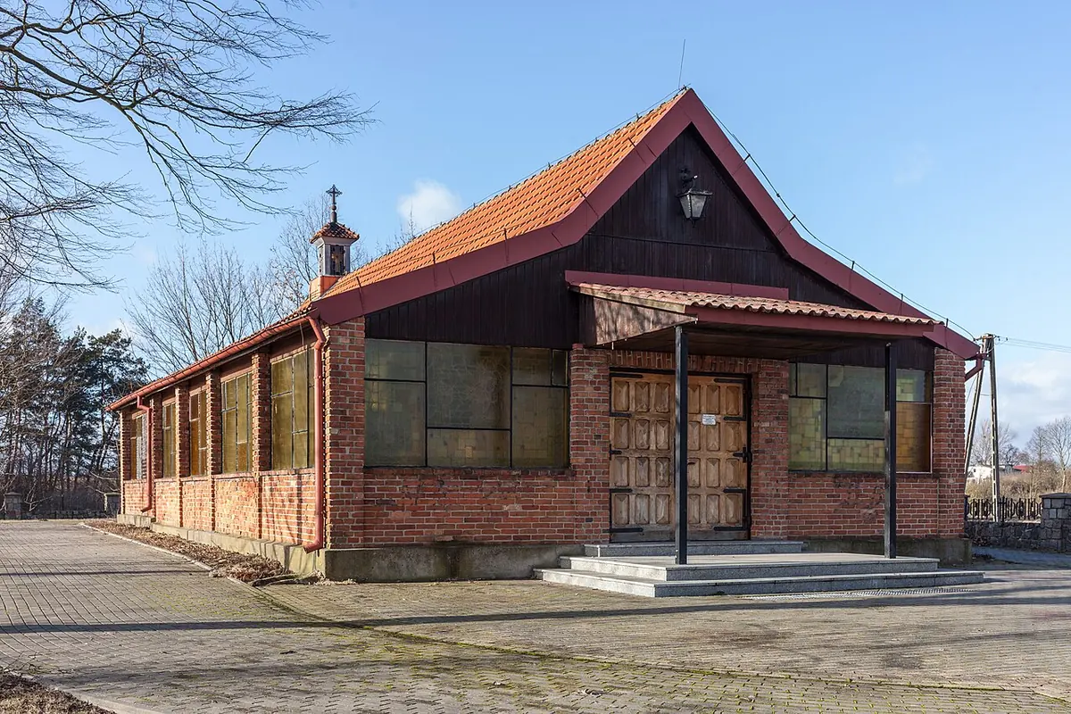 A long, low chapel of red brick with dark wooden gables and a small bell turret, set on a paved square under a clear winter sky