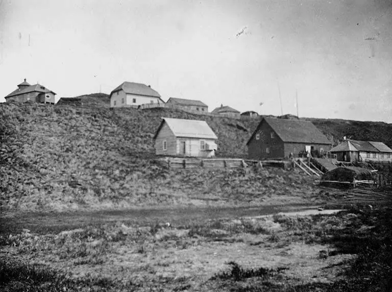Old black-and-white photograph of a few wooden houses and a trading post on a grassy bluff above a river beach in western Alaska