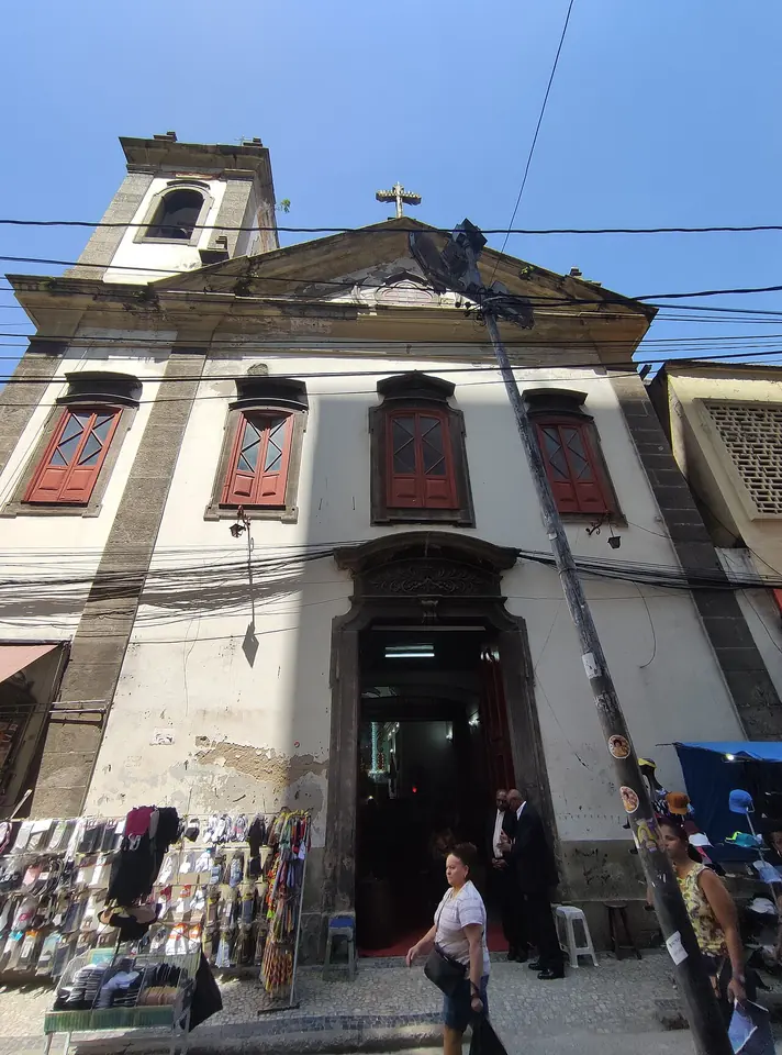 Whitewashed colonial church facade with a bell tower, red-shuttered windows and a cross, on a busy city street