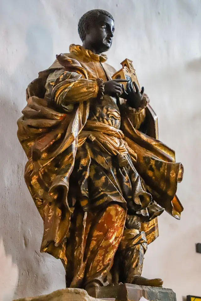 Gilded Baroque wooden statue of a dark-skinned saint in a flowing gold and brown robe and cloak, holding a small object in his hands