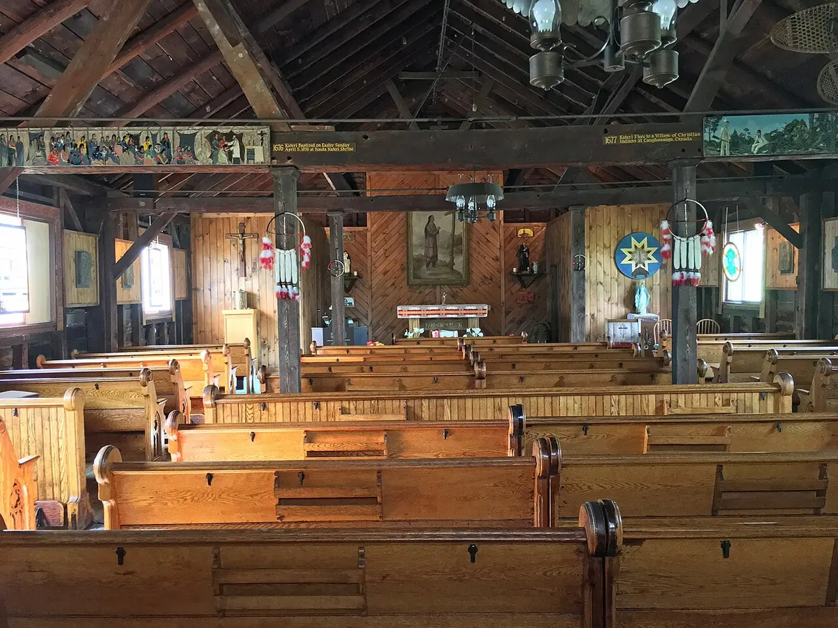 Interior of a wooden chapel with exposed roof beams, rows of pews and a painting of Kateri above the altar