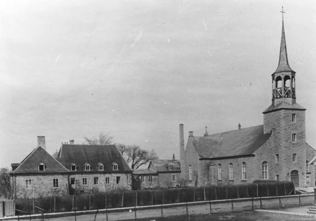 Old photograph of the stone Church of St. Francis Xavier at Kahnawake with its tall steeple, beside the long stone presbytery
