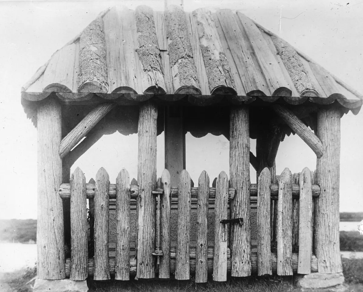 Black-and-white photograph of a rustic wooden shelter with a shingled roof and a picket fence, built over Kateri's grave near the St. Lawrence River