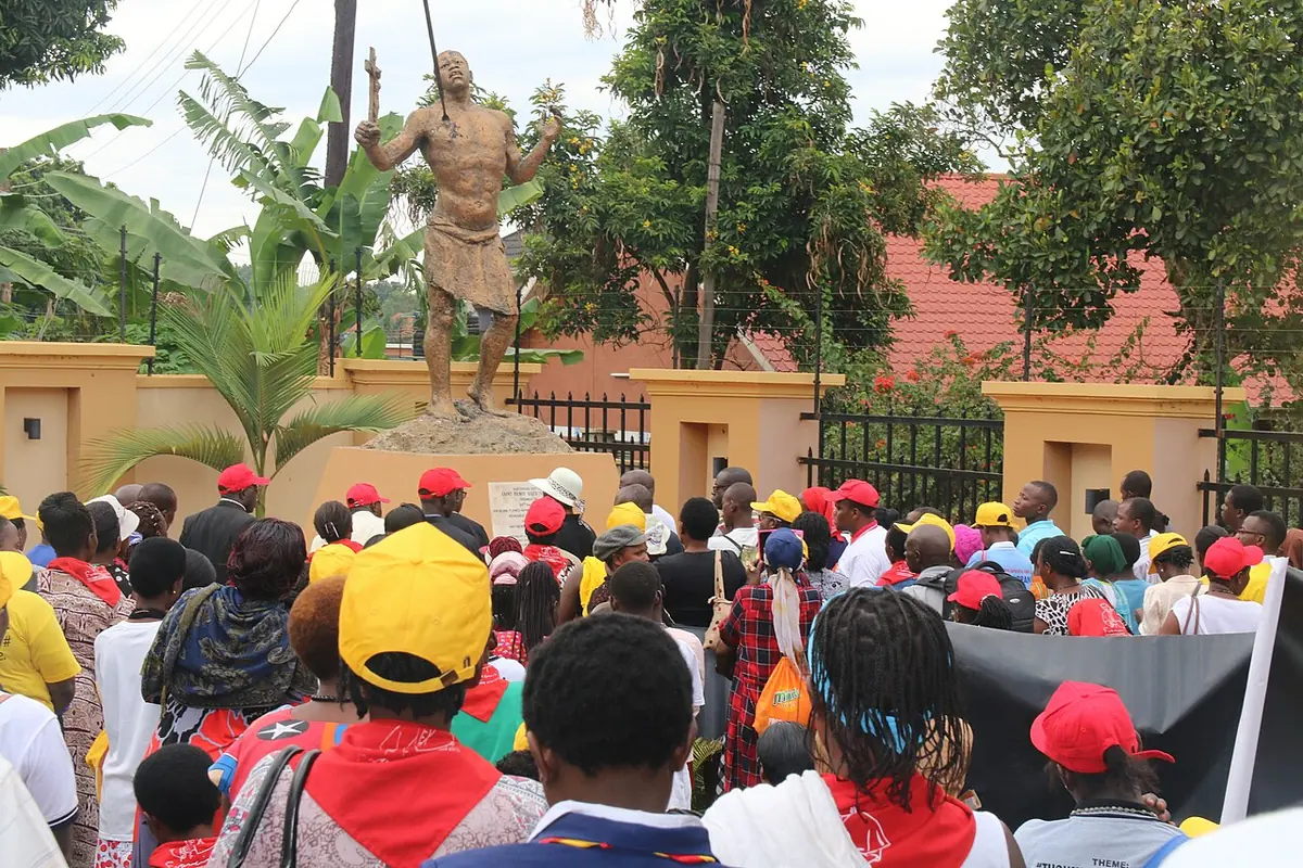 A crowd of pilgrims in red and yellow caps gathered before a bronze statue on a pedestal among trees at a shrine