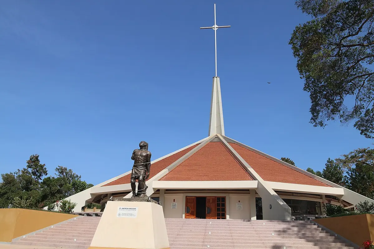 A modern church with a wide sloping red roof and a tall white spire, with a statue on a pedestal in front at the top of broad steps