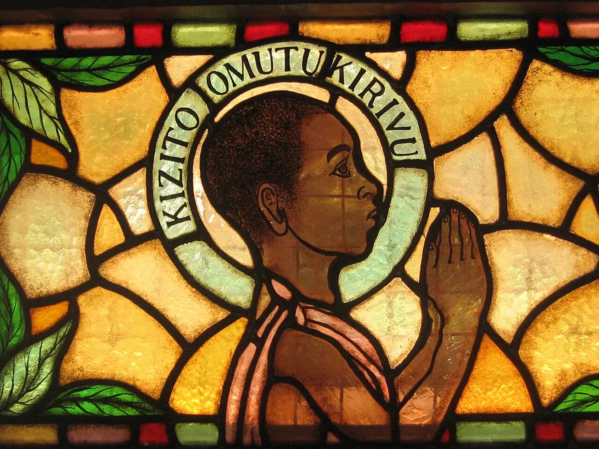 Stained glass of a young boy in profile with hands joined in prayer, a halo reading Kizito Omutukirivu, set among golden and green leaves