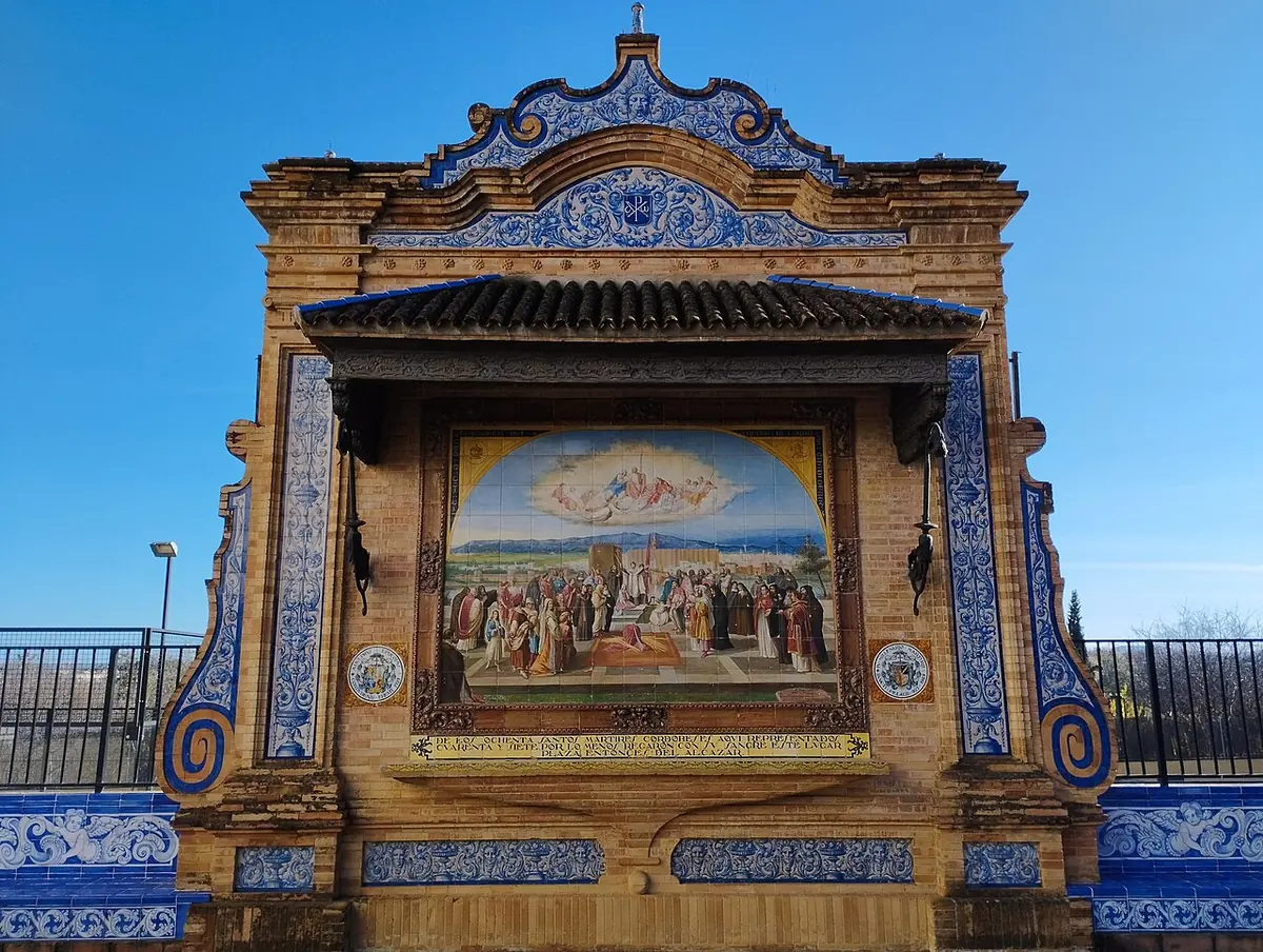 A painted tile mural of saints and a crowd beneath a glory of angels, framed by a brick and blue-and-white tile shrine under a small tiled roof
