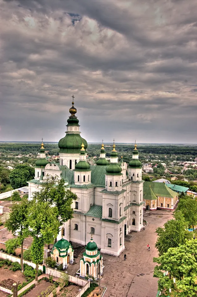 View down from a height onto a white cathedral with clusters of green domes and the monastery grounds, under a cloudy sky