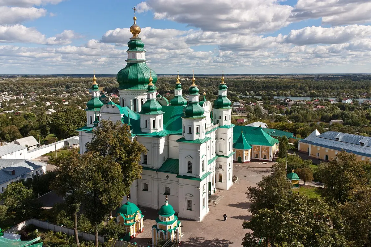 Aerial view of a large white Baroque cathedral with many green domes and gold crosses, inside a monastery courtyard, with the town of Chernihiv beyond