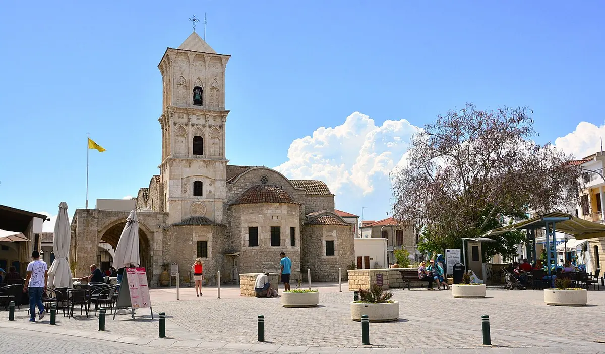 La iglesia de piedra de San Lázaro en Lárnaca, con su alto campanario de arcadas, en una plaza bajo un cielo azul con nubes blancas
