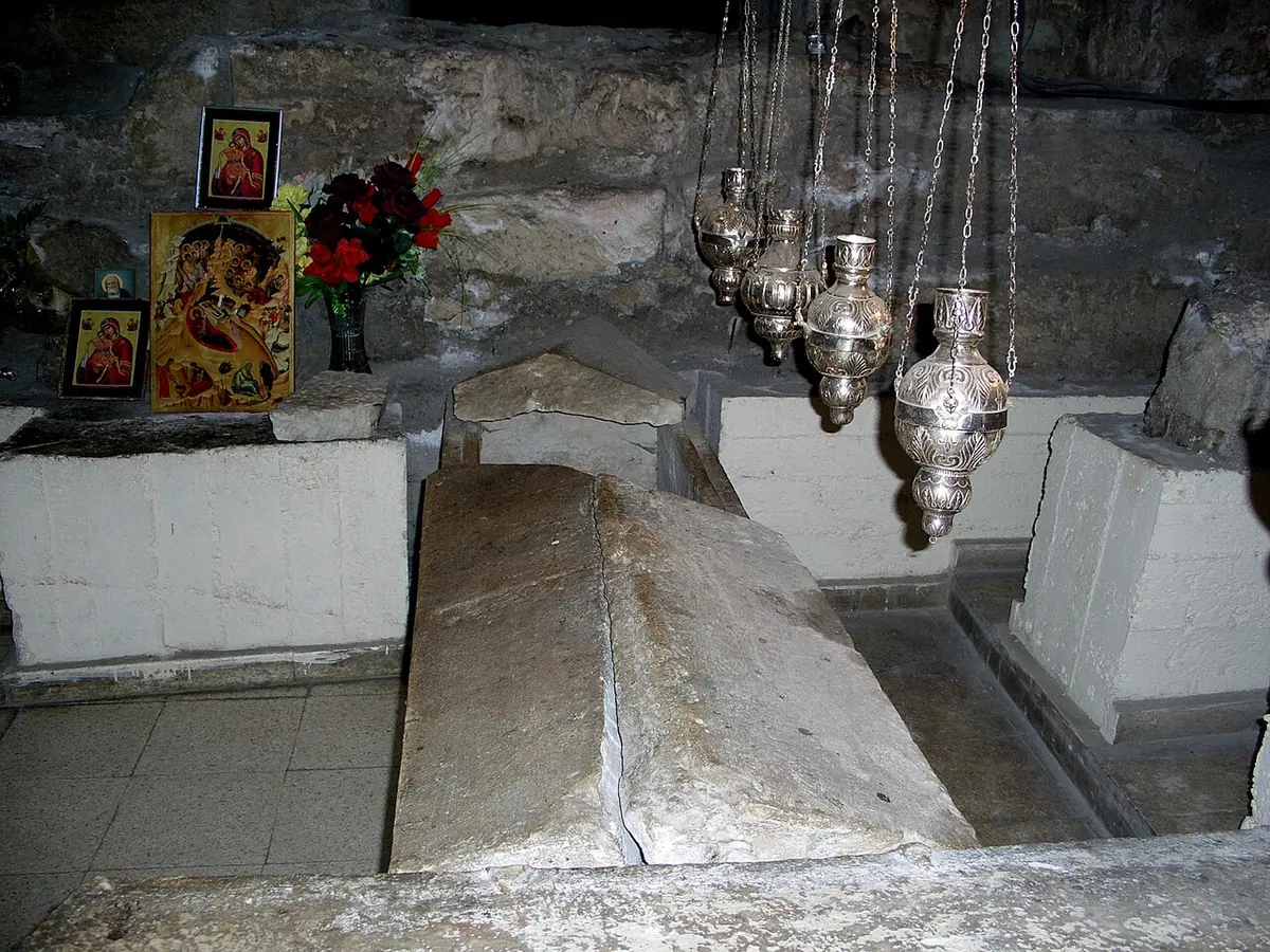 Sencillos sarcófagos de piedra en una cripta excavada en la roca, bajo lámparas de plata colgantes, con pequeños iconos y flores en una repisa