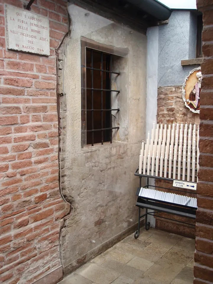 The outside wall of a small brick and plaster room with a barred window, a stone memorial plaque above it and racks of votive candles beside it