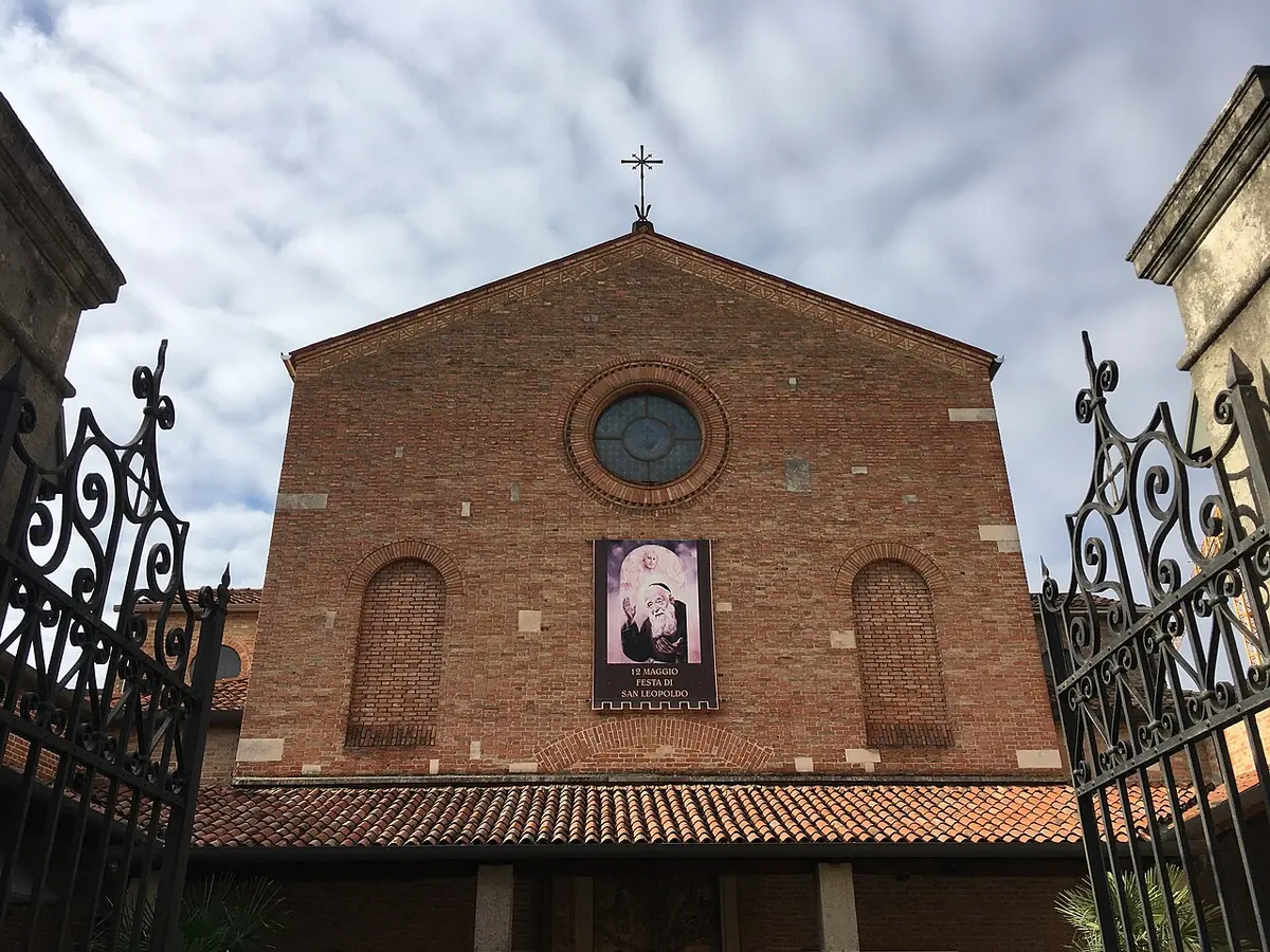 The brick front of a simple church with a round window and a large banner of the bearded friar, seen through open iron gates