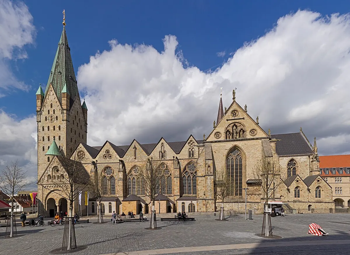 The long Gothic cathedral of Paderborn with its massive square Romanesque west tower and green spire, seen across an open square under a blue sky with clouds