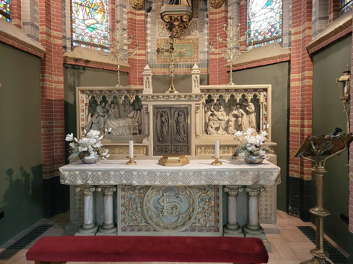 A carved white stone altar with relief panels and a small gilded tabernacle in a side chapel of red brick, with stained-glass windows above