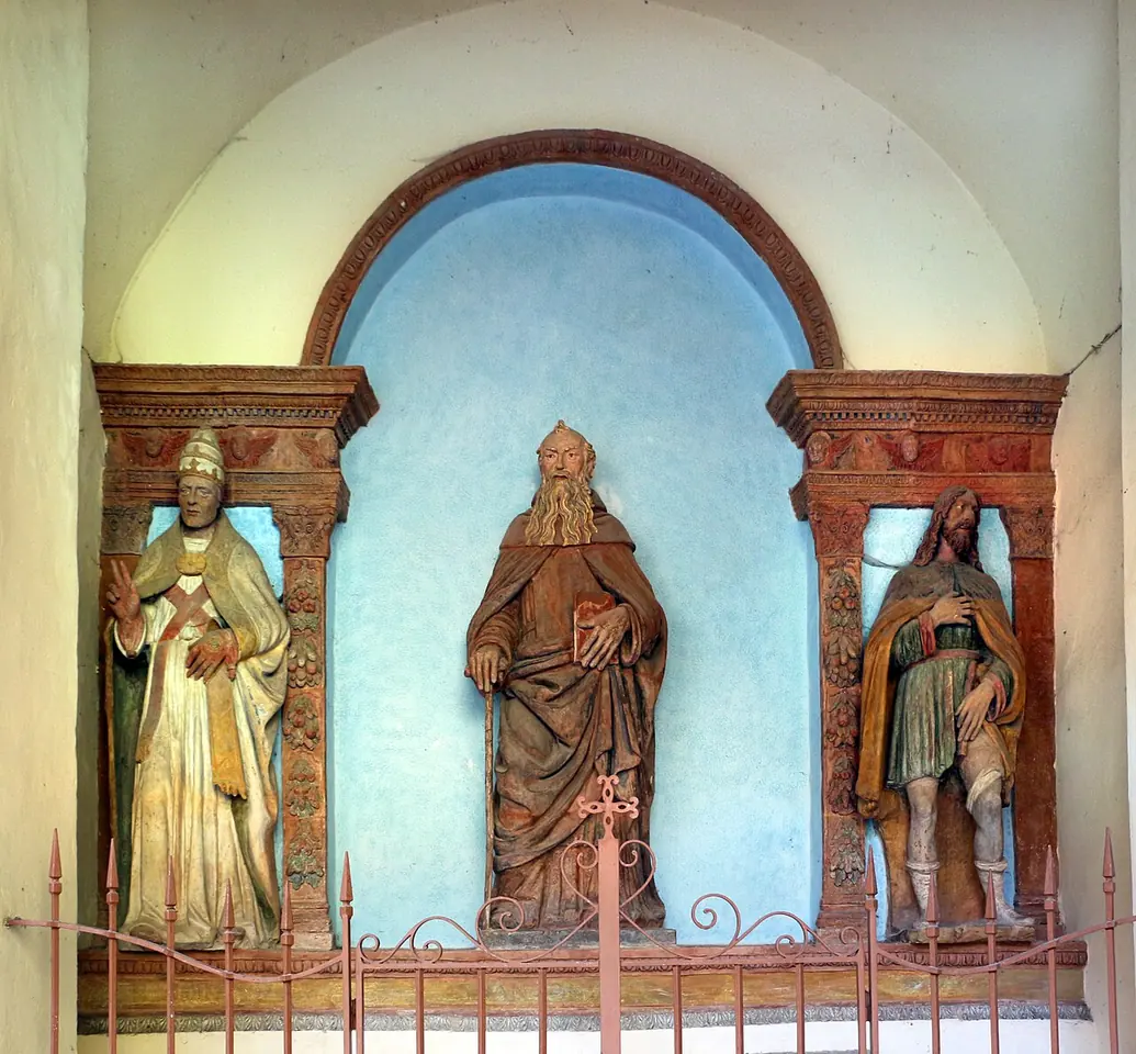 Three painted terracotta statues in an arched niche on a church wall, a pope in white with a tall tiara on the left, a bearded hermit in brown in the center and a pilgrim on the right