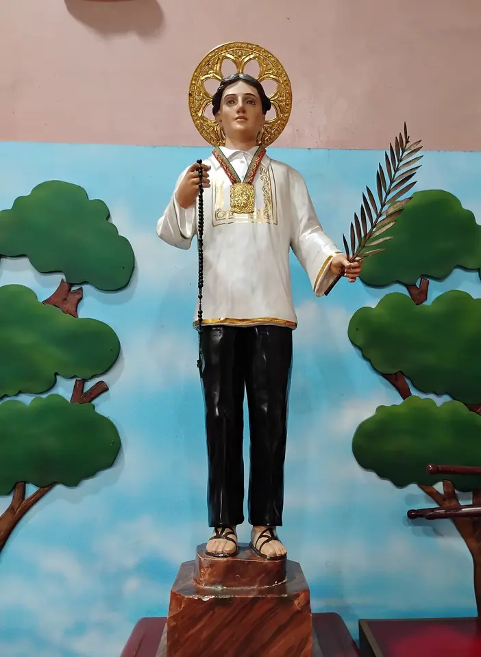 Painted statue of San Lorenzo Ruiz in a white embroidered shirt and black trousers, with a gold halo, holding a rosary and a palm, in front of a painted backdrop of trees