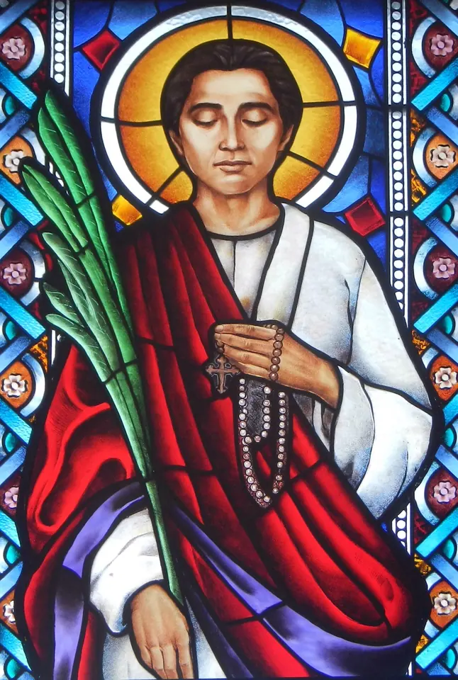 Stained-glass window of St. Lorenzo Ruiz with a gold halo, in a white shirt and red cloak, holding a rosary to his chest and a green palm in his other arm