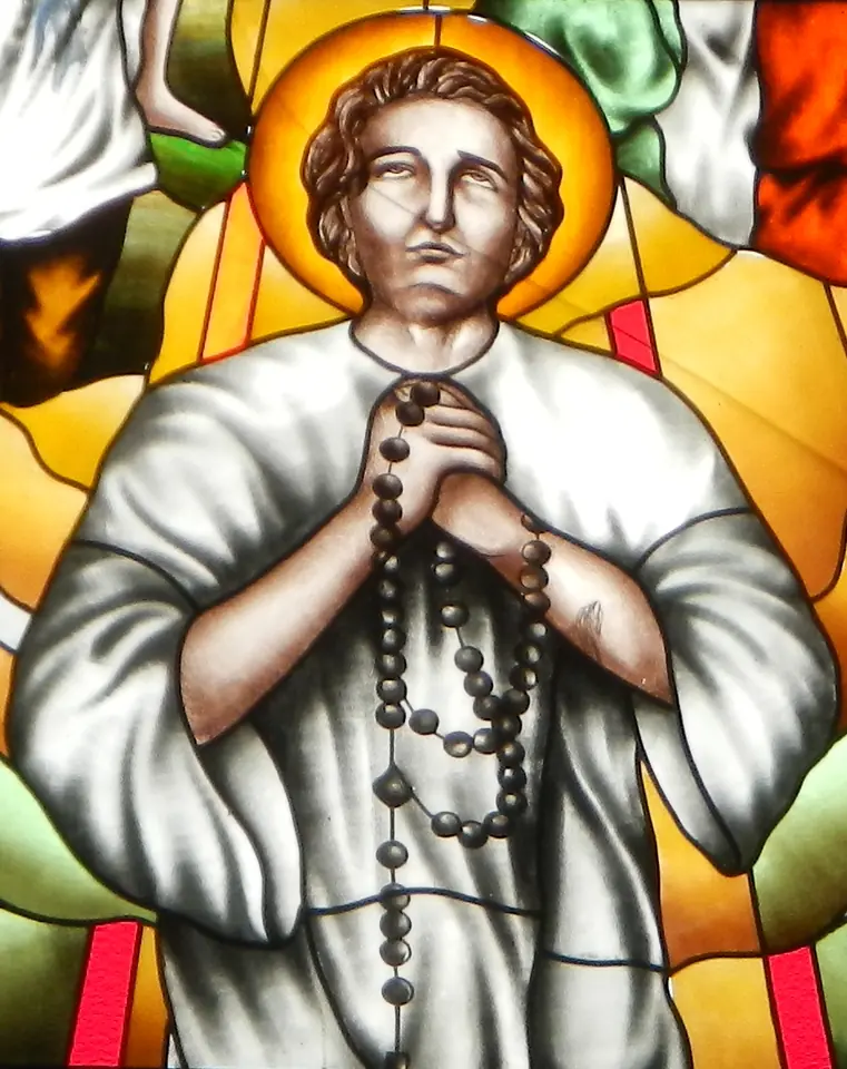 Stained-glass window of a young man with a gold halo and curly brown hair, in a white robe, praying with a long rosary wrapped around his folded hands